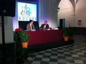 Setup for a MICE event in Malaga with a podium, professional lighting, sound equipment, a large screen displaying a presentation, and a projector in a modern conference venue.