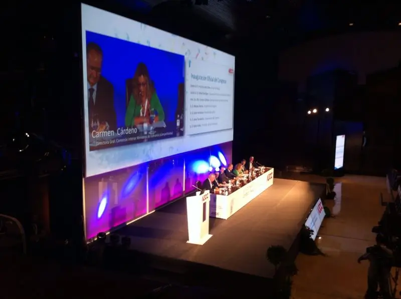 Setup for a MICE event in Malaga with a podium, professional lighting, sound equipment, a large screen displaying a presentation, and a projector in a modern conference venue.