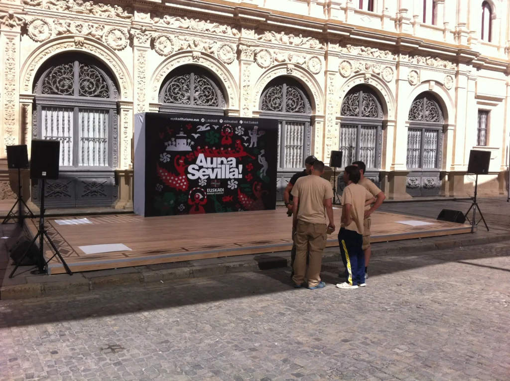 Technicians setting up a professional sound system on an outdoor stage in Malaga, with a decorative backdrop and speakers positioned on both sides.